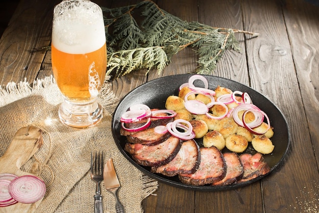 Almoço de presunto frito com batatas e cebolas em uma panela ao lado de um copo de cerveja