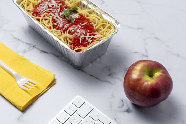 Almoço no local de trabalho espaguete caseiro e saudável com tomate e queijo na mesa de trabalho com teclado de computador. Comida em casa para o conceito de escritório. Comida para levar