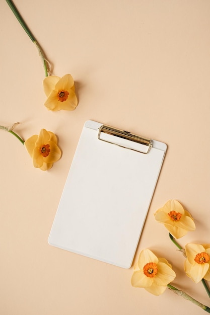 Almofada de tablet de prancheta de folha de papel de traçado de recorte em branco com flores de narciso de espaço de cópia de maquete em fundo de mesa de gengibre neutro Modelo de negócios mínimo de vista superior plana