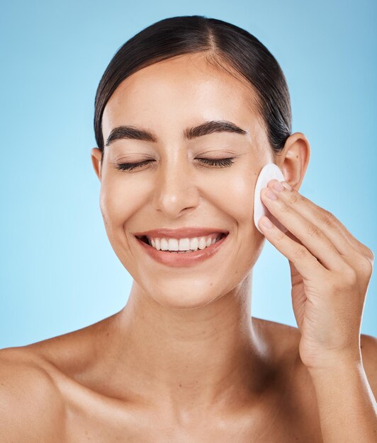 Foto almohadilla de algodón de belleza y mujer en un estudio quitando maquillaje cosmético o glamour facial cuidado de la piel y modelo femenino feliz con una rutina facial o de piel saludable aislada por un fondo azul