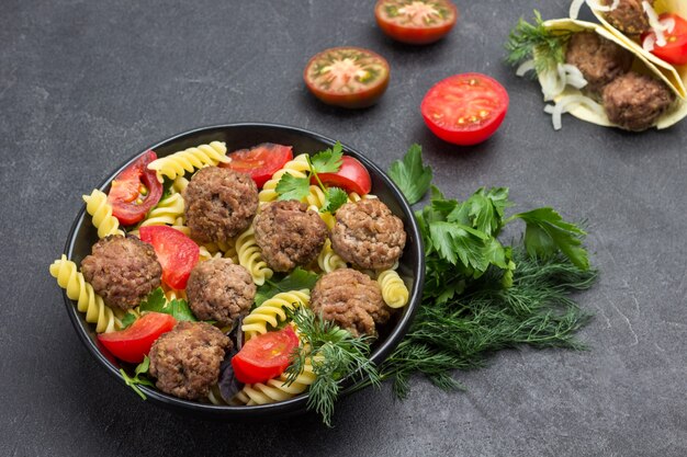 Almôndegas com tomate e macarrão em uma tigela preta. raminhos de endro e salsa. pita com almôndegas e cebolas. fundo preto. postura plana