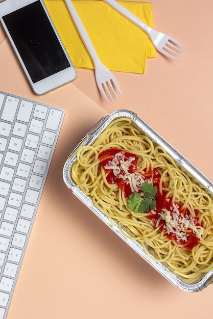 Almuerzo en el lugar de trabajo espaguetis caseros y saludables con tomate y queso en la mesa de trabajo con teclado de computadora. Comida casera por concepto de oficina. Comida para llevar