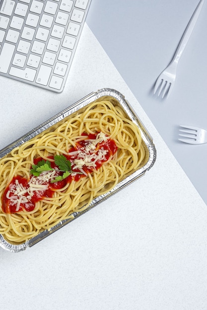 Almuerzo en el lugar de trabajo espaguetis caseros y saludables con tomate y queso en la mesa de trabajo con teclado de computadora. Comida casera por concepto de oficina. Comida para llevar