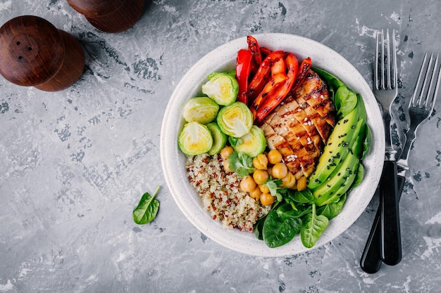 Almuerzo de tazón de buda vegetal saludable con pollo a la parrilla y quinoa espinacas aguacate coles de bruselas pimentón rojo y garbanzos sobre fondo gris Vista superior