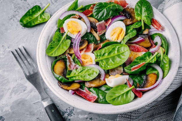 Almuerzo tazón de ensalada de espinacas con tocino, champiñones, huevos y cebollas rojas sobre fondo gris