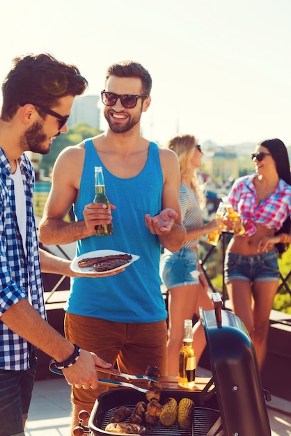 Almuerzo de verano perfecto. Dos hombres jóvenes alegres haciendo barbacoa y sonriendo mientras dos mujeres hablando entre sí en el fondo