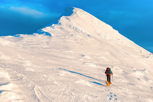 Alpinist mit Hund klettert auf einem schneebedeckten Berggipfel am Abend bei Sonnenuntergang