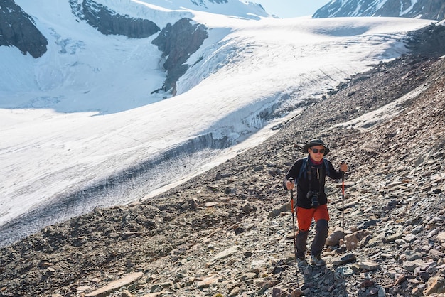 Alpinista com bastões de caminhada caminha perto da grande língua de geleira em altas montanhas Paisagem cênica com turista perto de geleira à luz do sol Homem com óculos escuros e câmera entre montanhas de neve em dia ensolarado