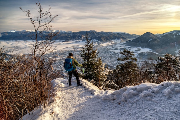 Alpinista com bastões de trekking e mochila olhando do topo da colina na paisagem de neve de inverno Colina Cebrat sobre a cidade Ruzomberok Eslováquia