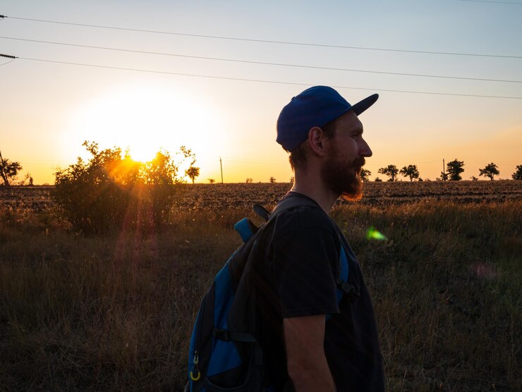 Foto alpinista com mochila em pé em uma colina e apreciando o pôr do sol homem barbudo em snapback caminhando conceito de natureza de viagem local