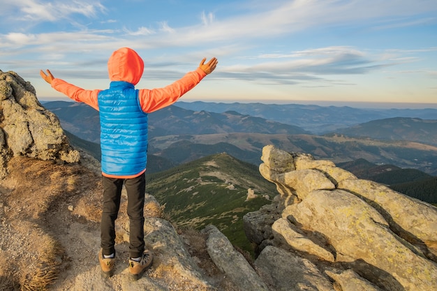Alpinista de menino jovem criança em pé com as mãos levantadas nas montanhas, apreciando a vista da paisagem de montanha incrível ao pôr do sol.