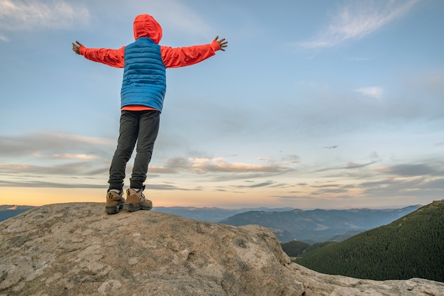 Alpinista de menino jovem criança em pé com as mãos levantadas nas montanhas, apreciando a vista da paisagem de montanha incrível ao pôr do sol.
