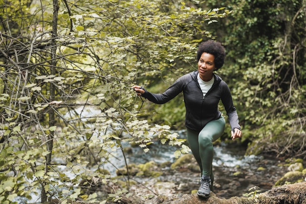 Alpinista de mulher afro-americana madura cruzando um riacho enquanto explorava as montanhas e desfrutava de treinamento ao ar livre.