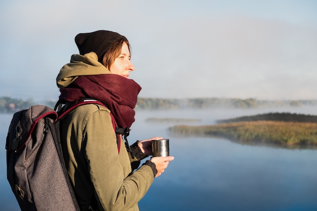 Alpinista de mulher gosta de belas paisagens naturais com um copo de bebida quente. Jovem fêmea tomando café e admirando o lindo rio coberto de névoa.