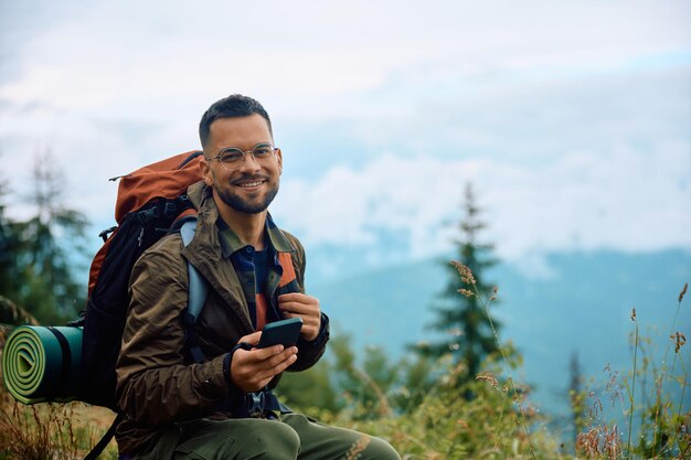 Alpinista feliz usando telefone celular enquanto relaxa na natureza e olhando para a câmera