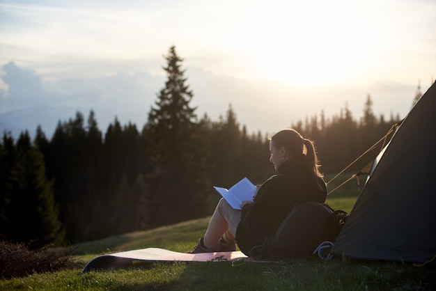 Alpinista jovem lendo um livro perto da tenda à noite ao pôr do sol