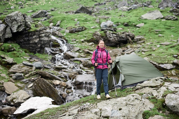 Alpinista morena feminina andando com bengalas nas mãos por uma encosta rochosa verde na frente de belas montanhas rochosas