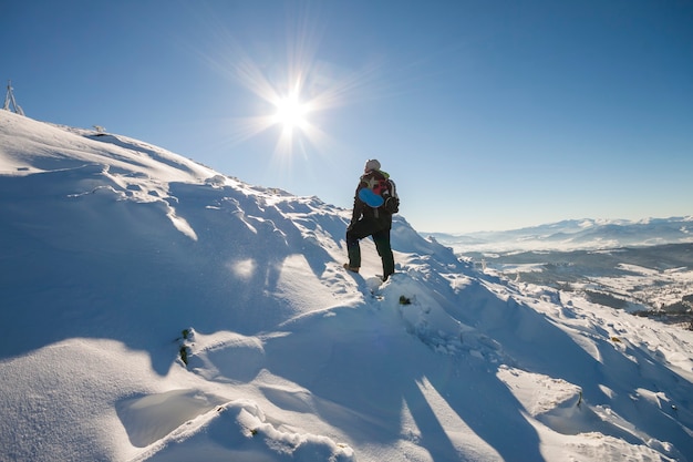 Alpinista turista alpinista em roupas de inverno com mochila escalando encosta de montanha rochosa íngreme perigosa coberto com neve profunda, raios de sol branco brilhando no fundo do espaço da cópia do céu azul brilhante.