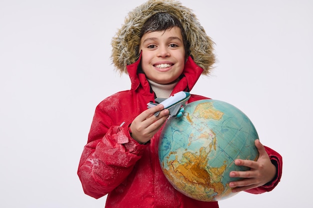 Alrededor del mundo concepto de viajes y turismo de invierno. Adorable niño de primaria en una cálida chaqueta de color rojo brillante con capucha sosteniendo un modelo de avión de juguete y un globo, imitando el vuelo alrededor de la Tierra