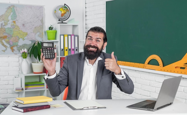 Alta pontuação de volta à escola educação informal feliz professor maduro mostrando resultado na calculadora homem barbudo brutal trabalho em sala de aula mostrar o polegar para cima preparar para o exame professor universitário na lição