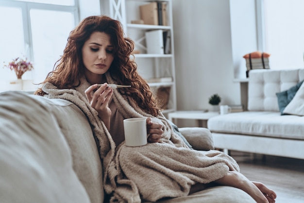 Alta temperatura. Mujeres jóvenes enfermas mirando el termómetro mientras está sentado en el sofá en casa