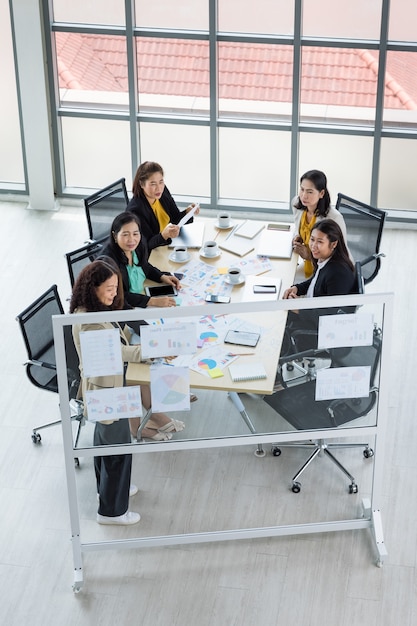 Alta visão de seis mulheres de negócios trabalhando e uma mulher apresentando gráficos e papéis de gráficos com tablets e laptops sobre a mesa. Conceito para reunião de negócios.