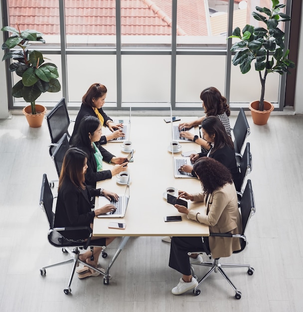 Alta vista de seis empresárias executivos sentados em cada cadeira, usando laptops e tablets na mesa de conferência de madeira na sala de reuniões no escritório. Conceito para reunião de negócios.