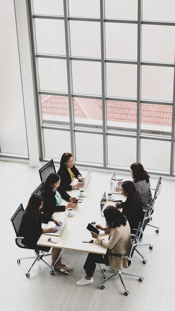 Alta vista de seis empresárias executivos sentados em cada cadeira, usando laptops e tablets na mesa de conferência de madeira na sala de reuniões no escritório. Conceito para reunião de negócios.