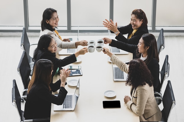 Alta vista de seis empresárias sentadas juntas ao redor da mesa de conferência de madeira, torcendo e tilintando na caneca de café junto com laptops e tablets na mesa no escritório. conceito para reunião de negócios.