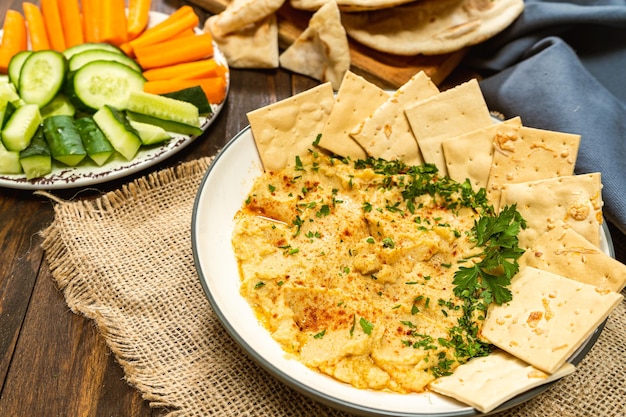 Alta vista de un tazón con Hummus casero con pan de pita y galletas saladas y en otro tazón zanahorias y otras verduras Concepto de alimentos frescos, saludables y naturales que se comen en casa