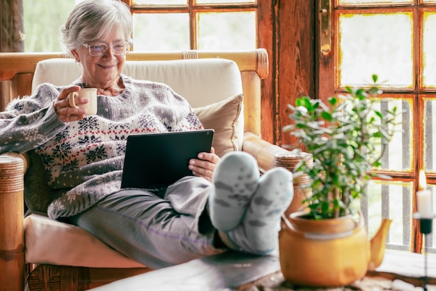 Alte ältere Frau, die zu Hause auf einem Sessel sitzt und ein digitales Tablet verwendet, das einen warmen Pullover und eine Brille trägt. Gemütliches Wohnzimmer, rustikale Holzfenster