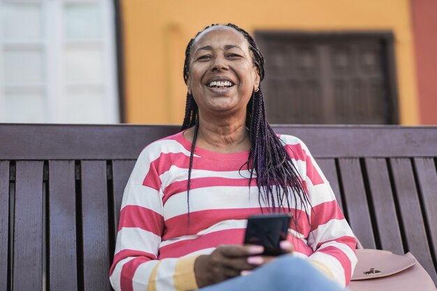 Alte afrikanische Frau, die auf einer Bank in der Stadt sitzt und vor der Kamera lächelt, während sie das Handy hält