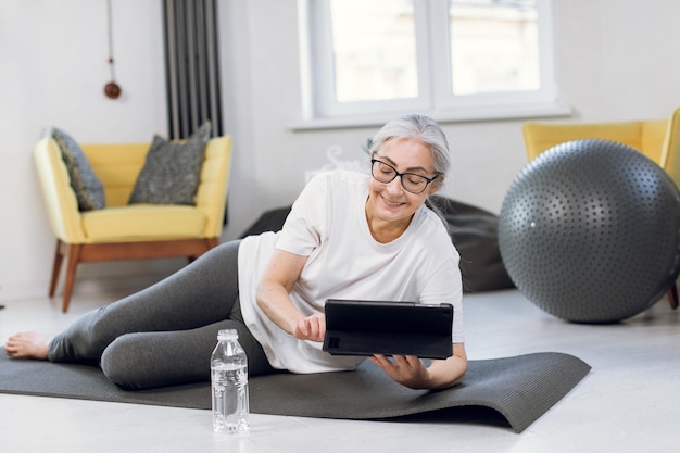 Alte Dame in Sportbekleidung mit Tablet, während sie auf Yogamatte liegt