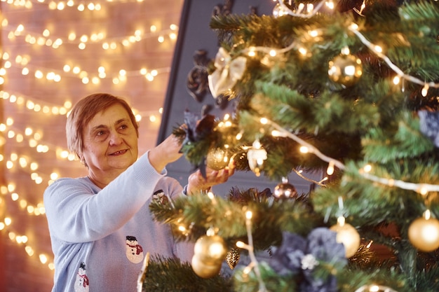 Alte Frau, die den Weihnachtsbaum drinnen im Zimmer schmückt.