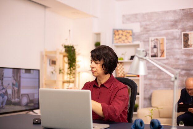 Alte Frau, die einen modernen Computer in ihrem Wohnzimmer benutzt, während ihr Mann im Hintergrund geht