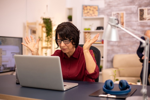 Alte Frau, die einen modernen Computer in ihrem Wohnzimmer benutzt, während ihr Mann im Hintergrund geht