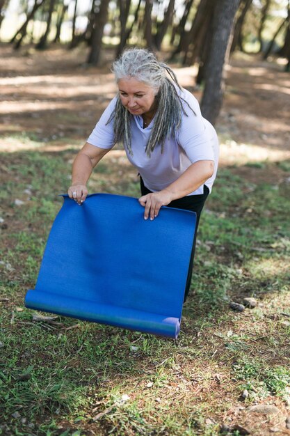 Alte Frau, die Yogamatte hält und sich darauf vorbereitet, Yoga oder Meditation im Freien im Park zu praktizieren, glücklich reif