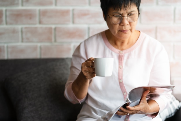 Alte Frau, die zu Hause ein Buch mit Tasse Kaffee liest