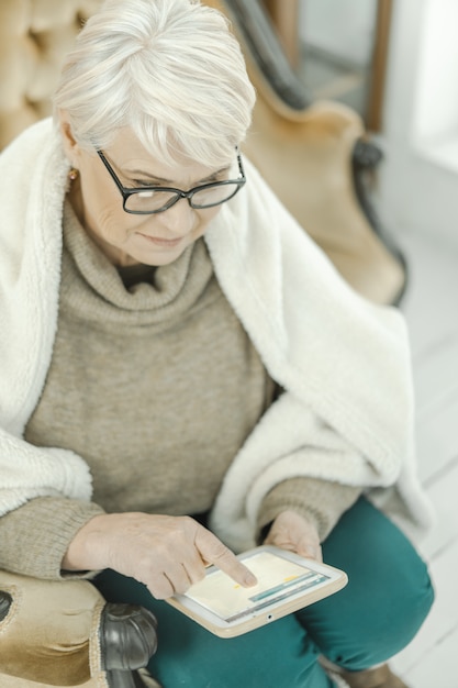 Alte Frau in Gläsern sitzt zu Hause auf dem Ledersofa mit einer Tablette in ihrer Hand