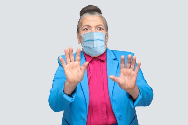 Alte Frau mit chirurgischer medizinischer Maske hat Angst, hat Angstgesicht. Großmutter mit hellblauem Anzug und rosafarbenem Hemd, das mit gesammelten grauen Haaren des Brötchens steht. Indoor-Studioaufnahme, isoliert auf grauem Hintergrund