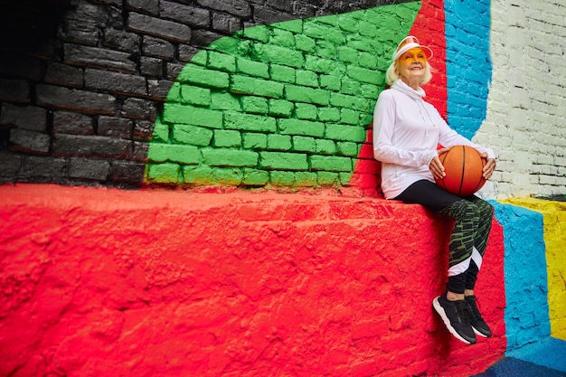 Alte, gesunde und fröhliche Dame mit Basketballball auf einem bunten Platz im Freien