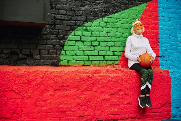 Foto alte, gesunde und fröhliche dame mit basketballball auf einem bunten platz im freien