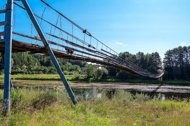 Alte hölzerne Hängebrücke am Fluss im sonnigen Sommertag. Natürliche Landschaft. Hund auf Brücke