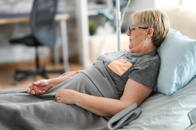 Alte Rentnerin in einem Pflegeheim, die auf dem Bett liegt und ihre Armtransfusion betrachtet.