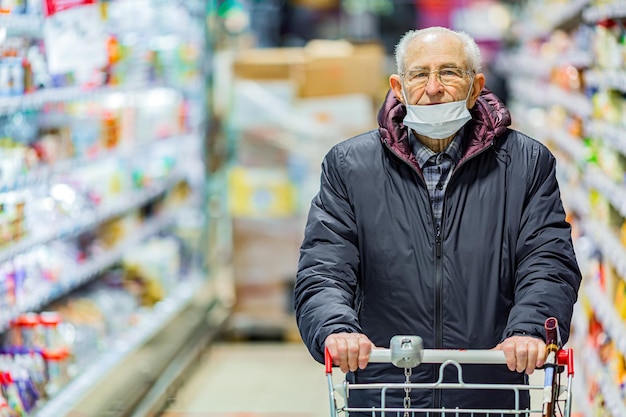 Alter älterer europäischer Mann mit schützender Gesichtsmaske, der Einkaufswagen im Supermarkt schiebt. Einkaufen während des COVID-19-Konzepts.