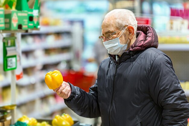 Alter älterer europäischer Mann mit schützender Gesichtsmaske mit Blick auf gelben Paprikapfeffer im Supermarkt. Einkaufen während des COVID-19-Konzepts.