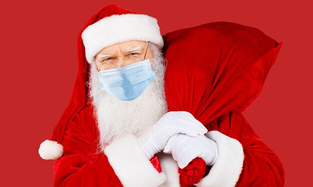 Alter bärtiger glücklicher Weihnachtsmann trägt Atemschutzmaske auf rotem Hintergrund