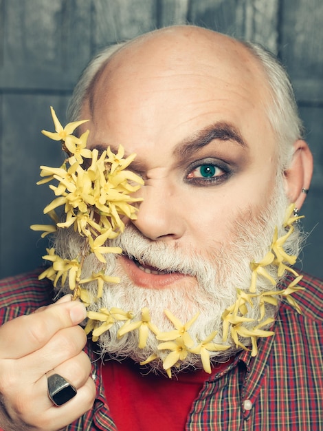 Alter bärtiger Mann mit gelben Blumen im langen grauen Bart auf lächelndem glücklichem Gesicht im rot karierten Hemd auf hölzernem Hintergrund