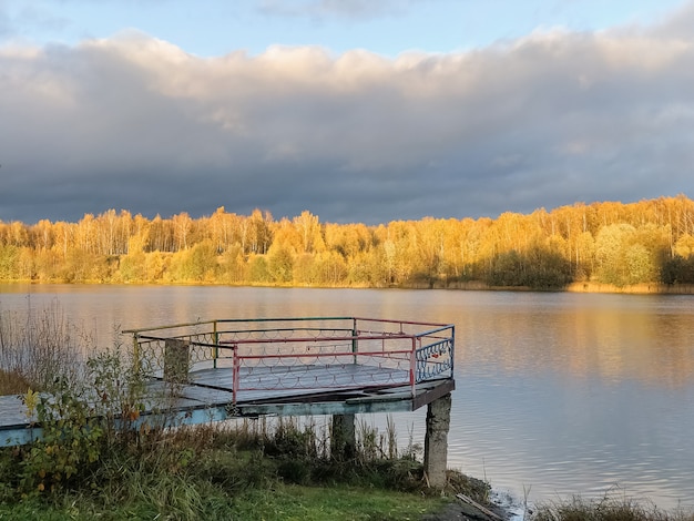 Alter Holzsteg über stillem Seewasser mit herbstlichen gelben Waldbäumen im Horizont