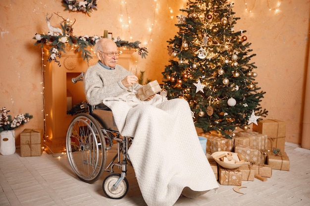 Alter Mann auf einem Rollstuhl, der nahe Weihnachtsbaum sitzt und eine Geschenkbox hält